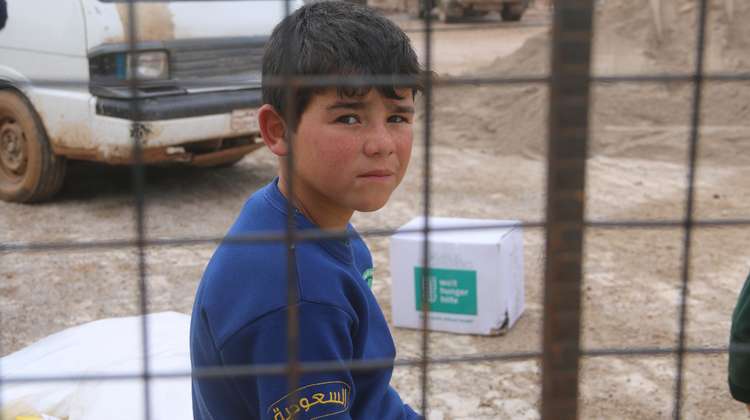 Ein Junge sitzt hinter einem Gitter und schaut in die Kamera. Im Hintergrund sieht man ein Welthungerhilfe-Paket.