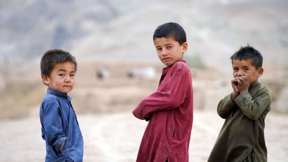Drei Kinder in Afghanistan schauen in die Kamera. Im Hintergrund ist karge Landschaft zu sehen.