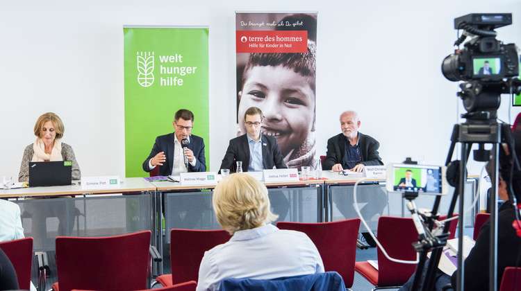 Vier Menschen sitzen in einer Reihe an einem Tisch im Hintergrund das Logo von der Welthungerhilfe und das Logo von terre des hommes.