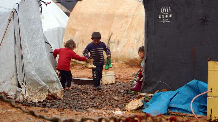 Drei Kinder mit Gummistiefeln zwischen Zelten in einem matschigen Flüchtlingscamp. 