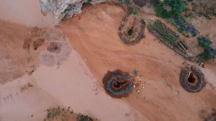 Luftaufnahme von einem ausgetrockneten Flussbett mit rötlicher Erde. Rechts sind drei Wasserlöcher zu sehen, um die herum etwas Grün wächst. 