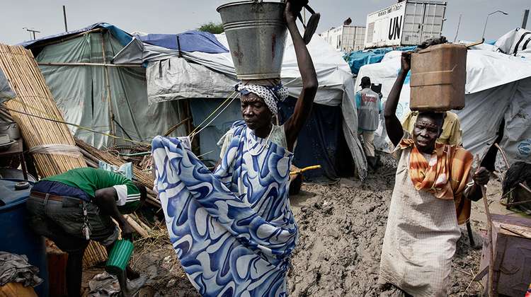 Ihre Hilfe für Menschen im Südsudan. Bildbeschreibung: Frauen schleppen Behälter auf dem Kopf, im Hintergrund sind viele Zelte zu sehen.
