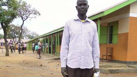 Akoy steht auf dem Schulhof, hinter ihm befindet sich das Schulgebäude.