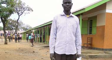 Akoy steht auf dem Schulhof, hinter ihm befindet sich das Schulgebäude.
