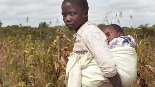 Spenden für Simbabwe - Bild: Ein junges Mädchen mit einem Baby auf dem Rücken schaut in die Kamera.