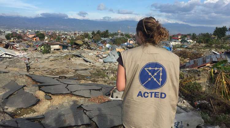 Eine Frau steht mit dem Rücken zur Kamera und schaut auf eine Reihe zerstörter Häuser.