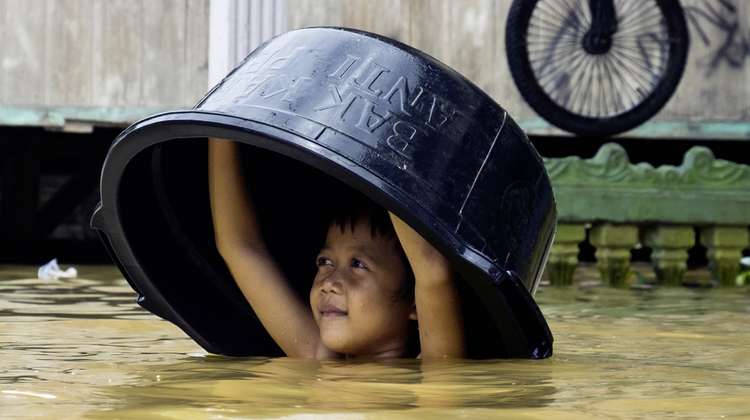 Spenden Indonesien. Bildbeschreibung: Ein Junge mit einer Schüssel über dem Kopf watet durch Wasser.