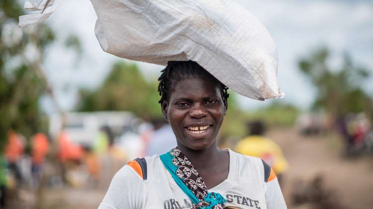 Eine Frau trägt ein Hilfsgüter-Paket auf dem Kopf und lächelt in die Kamera.