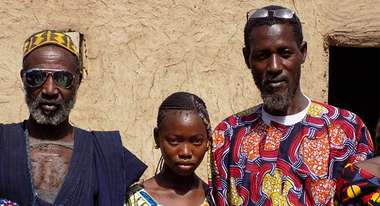Massiga Diawara vor einer Hausmauer in seinem Dorf einem Mädchen und einem älteren Mann © Schwenk
