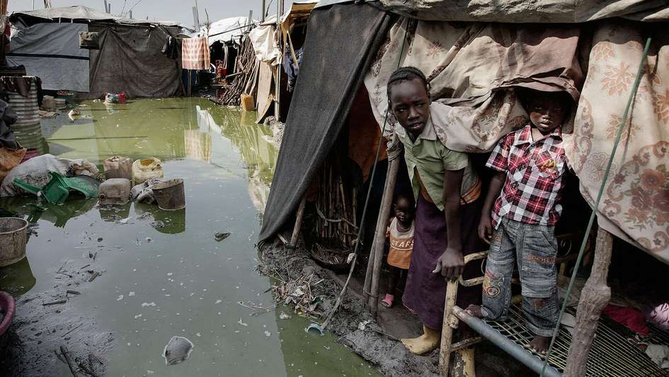 Flüchtlingslager im Südsudan: Knietiefes Wasser umgibt ein Zelt, aus dem drei Menschen herausschauen