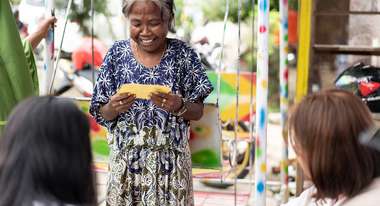 Eine ältere Frau hält einen gelben Zettel in der Hand, schaut auf ihn und lächelt.