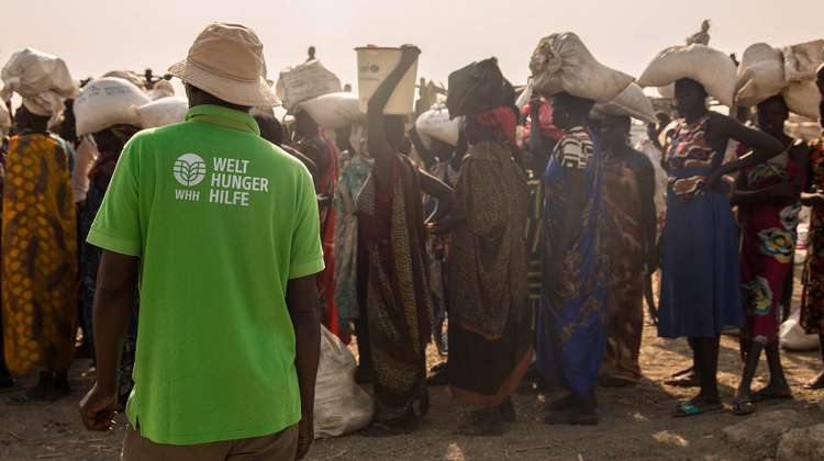 Mitarbeiter der Welthungerhilfe in einem grünen T-Shirt mit dem neu gestalteten Welthungerhilfe-Logo. Im Hintergrund sind Menschen zu sehen, die Dinge auf ihren Köpfen tragen.