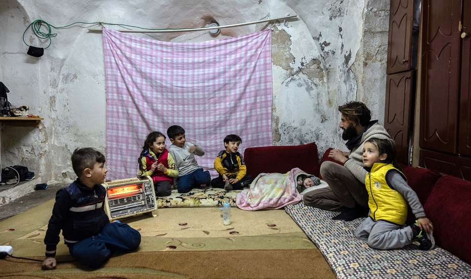 Ein Familienvater und fünf Kinder sitzen im Wohnraum eines alten Hauses auf Sitzkissen und Teppichen vor einem Heizstrahler und reden miteinander.